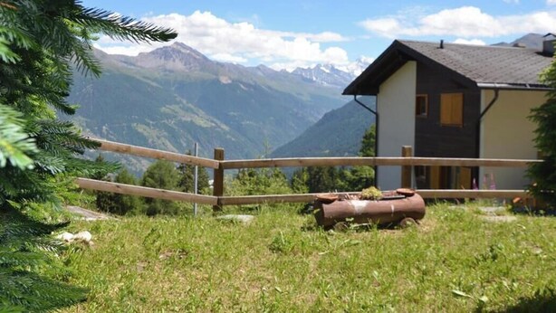 A mountain cabin with a wooden fence, a small shed, and a view of snow-capped mountains.