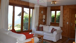 A cozy living room with wooden walls, a white sofa, a wooden coffee table, and large windows offering a view of greenery and mountains.