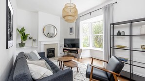 Un salon moderne avec un canapé gris, une table basse en bois et un fauteuil en cuir noir. On y trouve une bibliothèque, une cheminée et une grande fenêtre avec des rideaux.