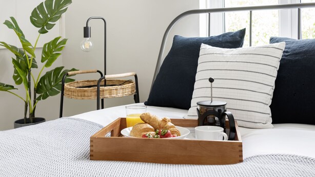Un lit bien fait, orné d’oreillers bleu foncé, une assiette avec des croissants, des fraises et un verre de jus d’orange.