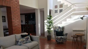 Un salon comprenant un canapé, un fauteuil à motifs, un tapis vert, une plante en pot et un escalier orné de carreaux décoratifs.