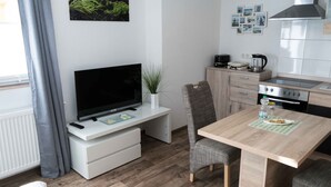 Espaço compacto para viver com televisão em suporte branco, pequena mesa de jantar com uma cadeira, cozinha com fogão e forno, e uma planta no balcão.
