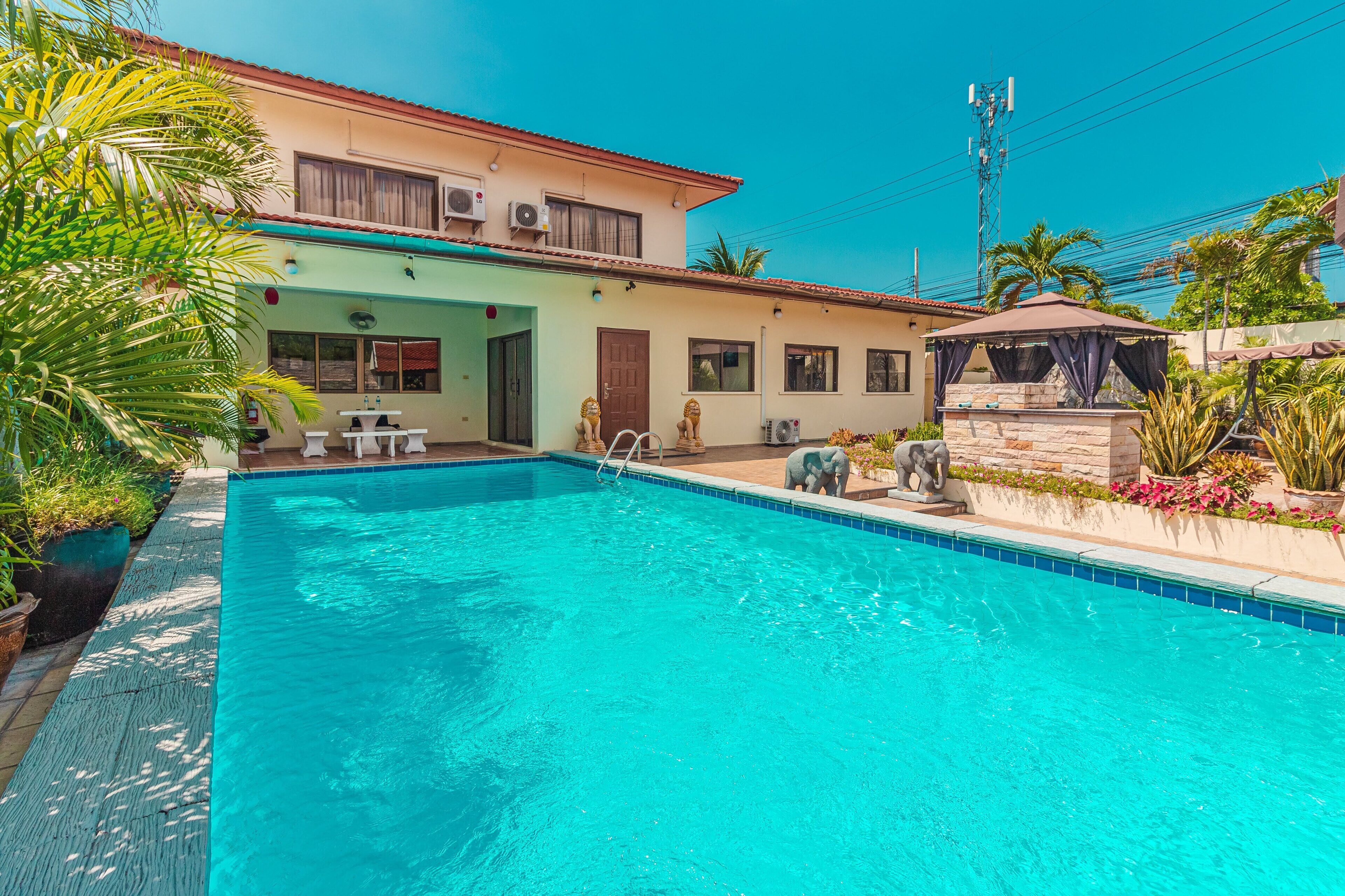 A swimming pool with a clear blue water, surrounded by a house with a red roof, a covered patio, and a stone wall with statues.