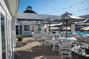 Eine Terrasse mit Sitzgelegenheiten und Sonnenschirmen, ein Pool und ein Gebäude mit großen Fenstern.
