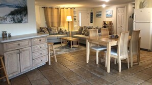 Ein helles, offenes Wohn- und Esszimmer mit Küche, einer Sofagarnitur mit gemusterten Kissen, einem Esstisch mit Stühlen und einem Kühlschrank.