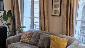A living room with a grey sofa, patterned cushions, a framed picture on the wall, and a potted plant.