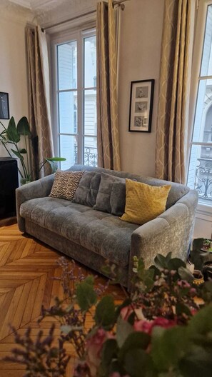 Una sala de estar con un sofá gris, cojines estampados, un cuadro en la pared y una planta en maceta.