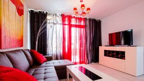 A modern living room with a sectional sofa, a white coffee table, a TV on a white cabinet, and a large window with red curtains.