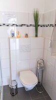 Un baño pequeño con inodoro, un cubo de basura, ducha y un estante con artículos de aseo.