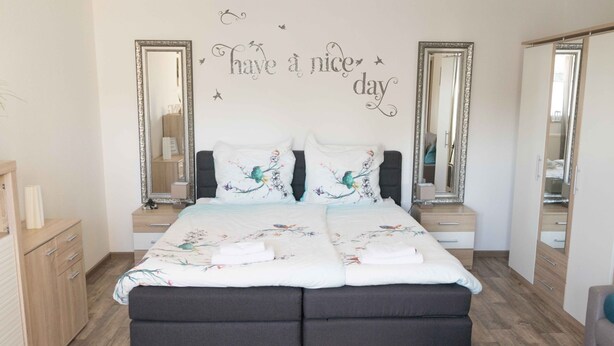 A bedroom with a double bed, two nightstands, two large mirrors, and a decorative wall sign. - Ferienwohnung Anke-Apartement 3c (Heinsberg)
