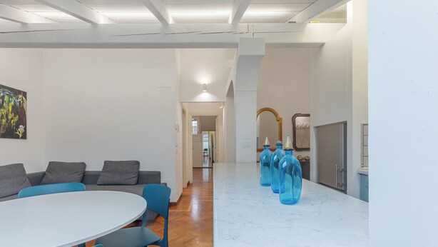 A modern living room with a round table and blue chairs, a sofa with gray cushions, and a kitchen area with a marble countertop and blue glass bottles. - Pratello Elegante e Raffinata Mansarda (Bologna)