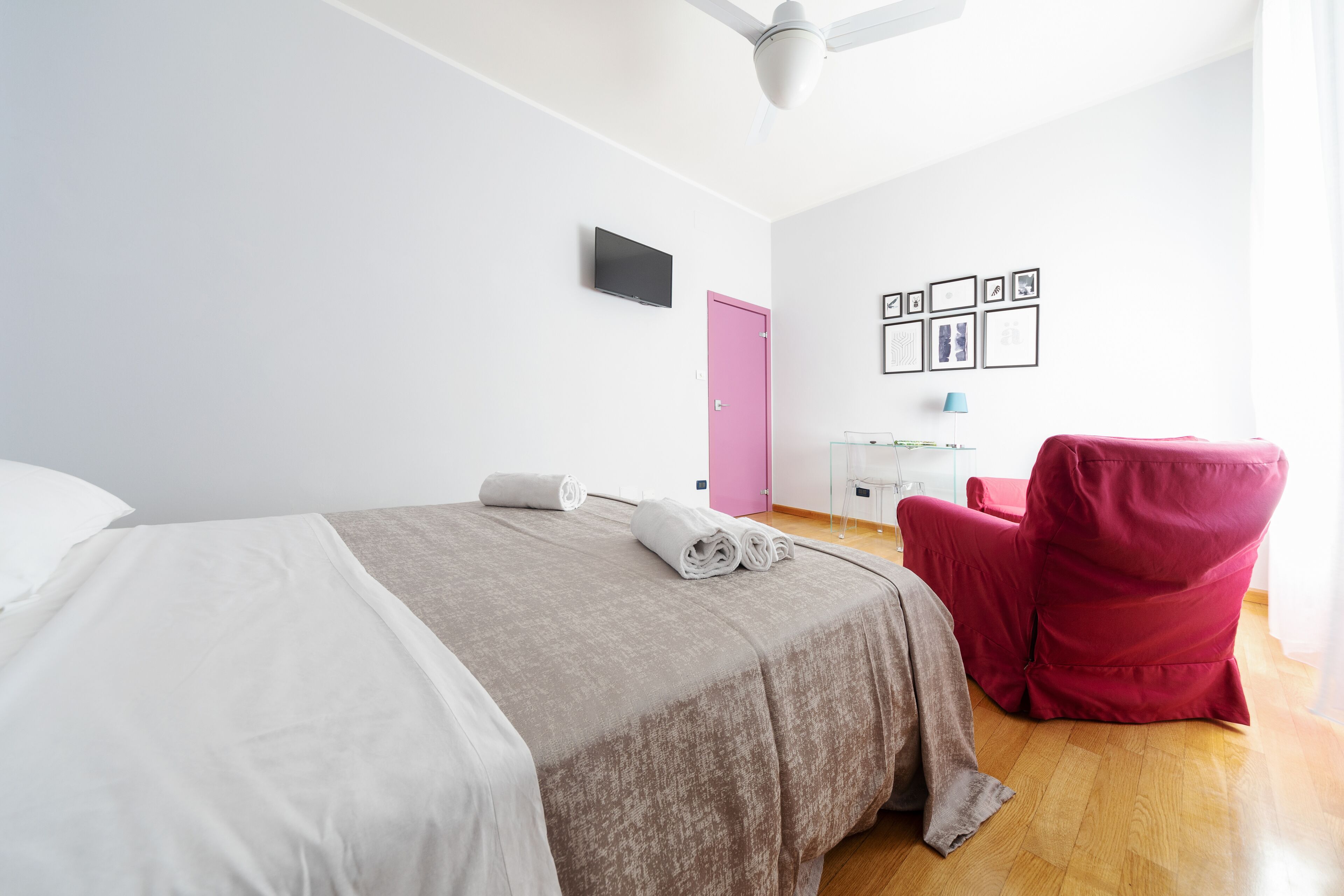 Habitación de hotel con cama, una puerta rosa, escritorio con lámpara y una silla roja.