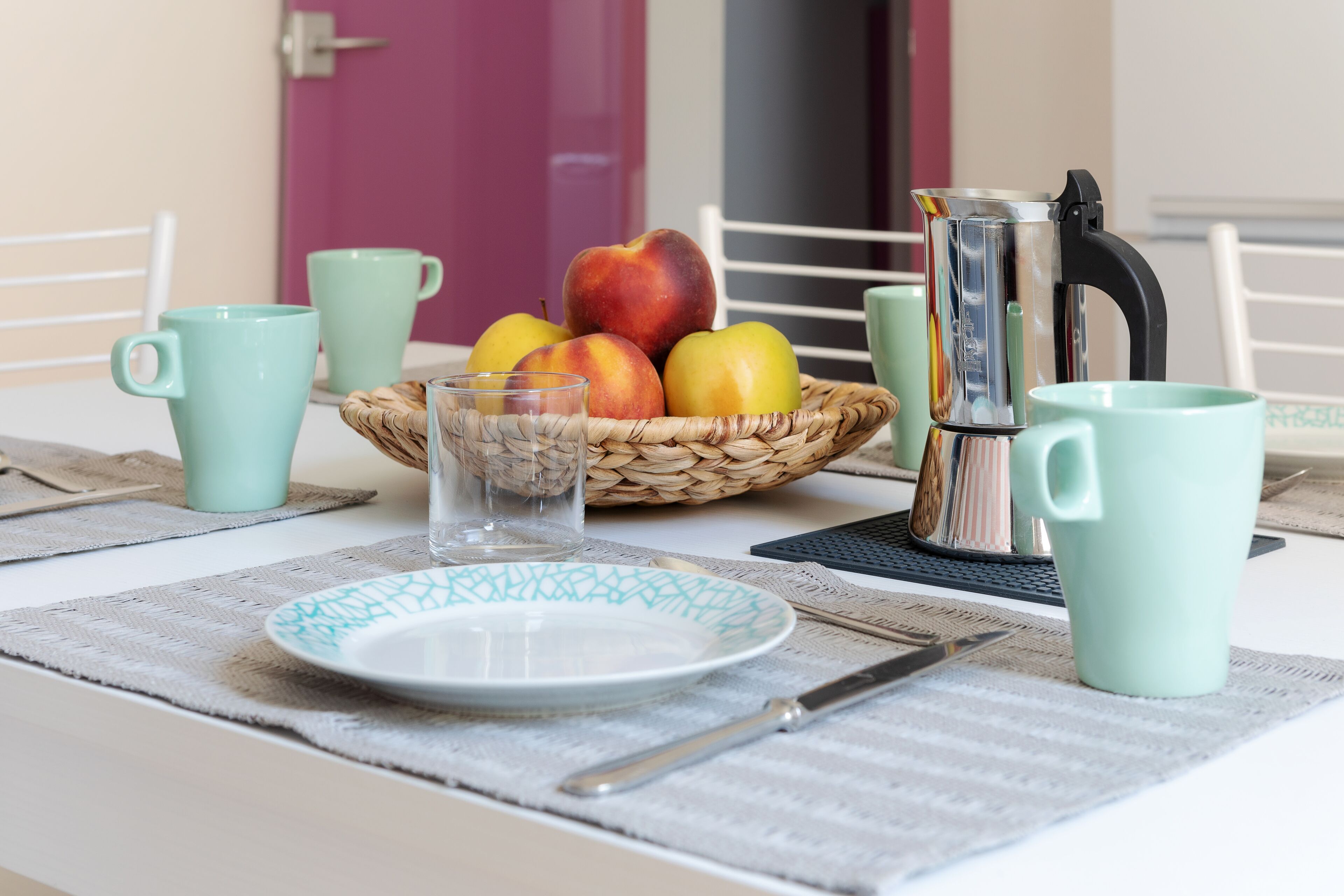 Uma mesa com disposição para o café da manhã, incluindo um prato, talheres, um copo, uma cesta de frutas e uma cafeteira.