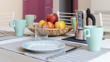 Uma mesa com disposição para o café da manhã, incluindo um prato, talheres, um copo, uma cesta de frutas e uma cafeteira.