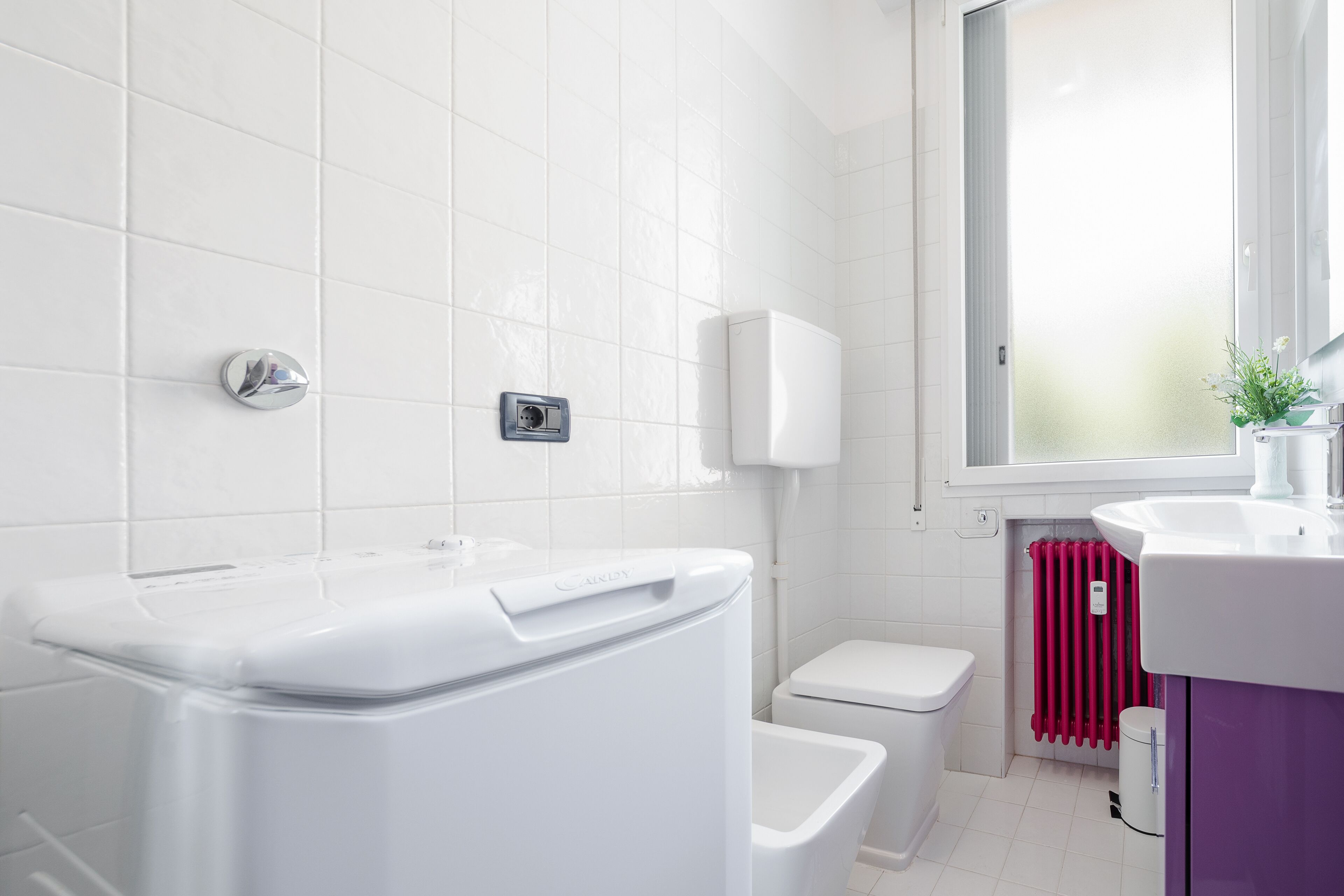 Ein modernes Badezimmer mit weißen Fliesen, einer weißen Toilette, einem weißen Bidet, einem weißen Waschbecken und einem Fenster mit Aussicht.