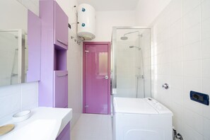 A bathroom with a white sink, a washing machine, a glass shower enclosure, and a purple door. - Sant'Orsola Colorful Huge Apartment (Bologna)