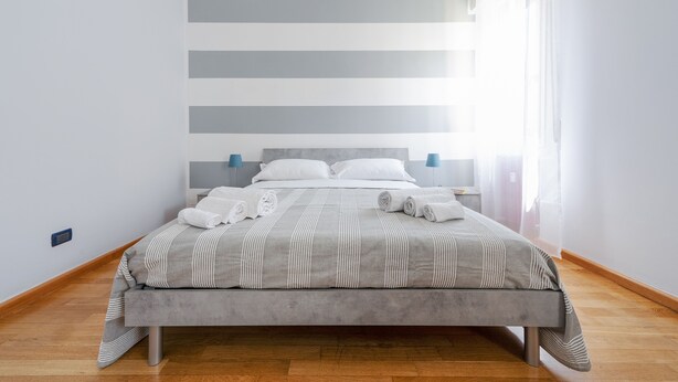 A modern bedroom with a grey-striped bedspread, a wooden floor, and a large window with sheer curtains. - Sant'Orsola Colorful Huge Apartment (Bologna)