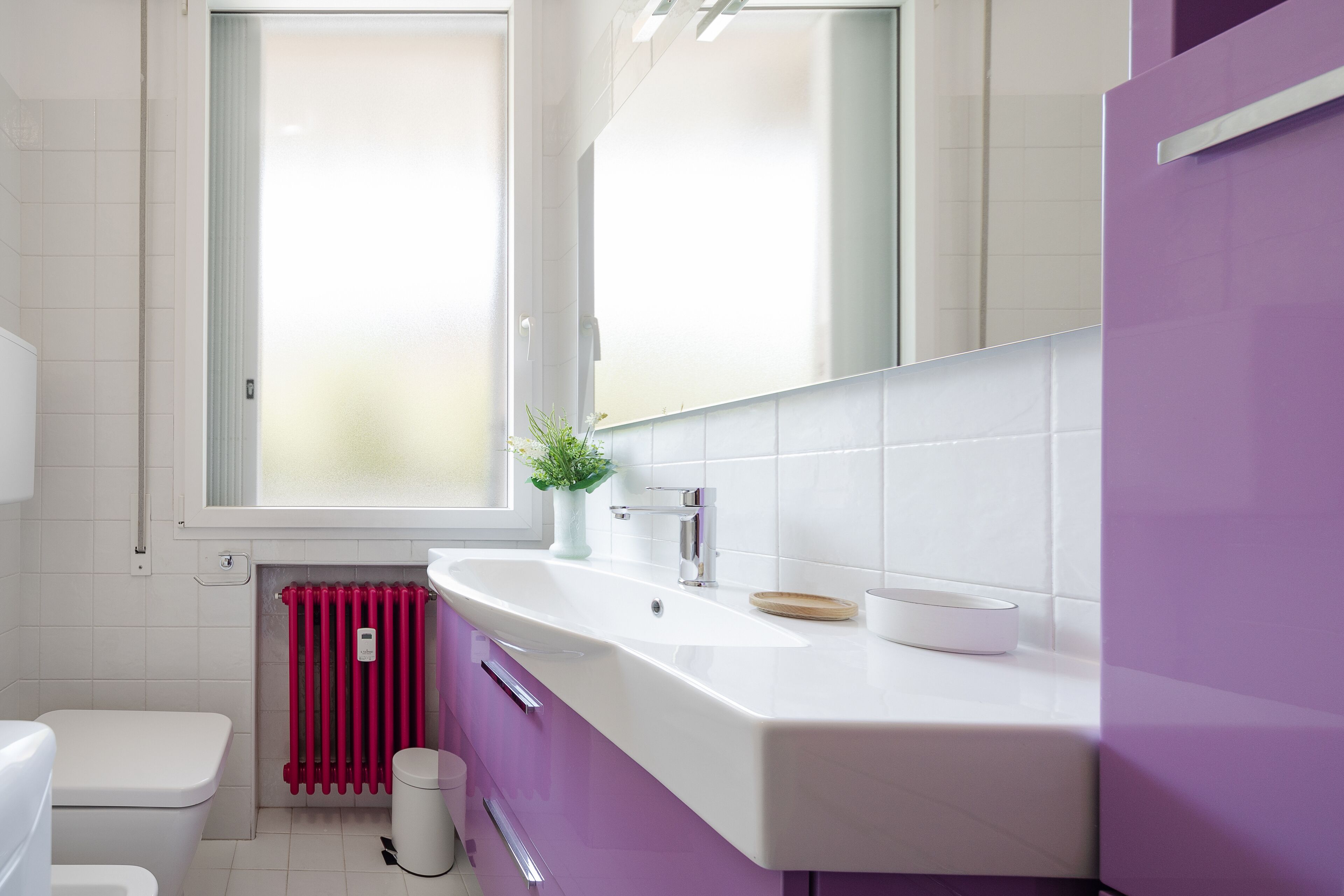 A modern bathroom with a white sink, purple cabinets, a white toilet, and a red radiator.