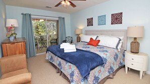 A bedroom with a bed, a ceiling fan, a nightstand, a chair, and a view of the outdoors. - TB 62C Updated 3 Bedroom 2 Bath Condo in True Blue Golf Community (Pawleys Island)