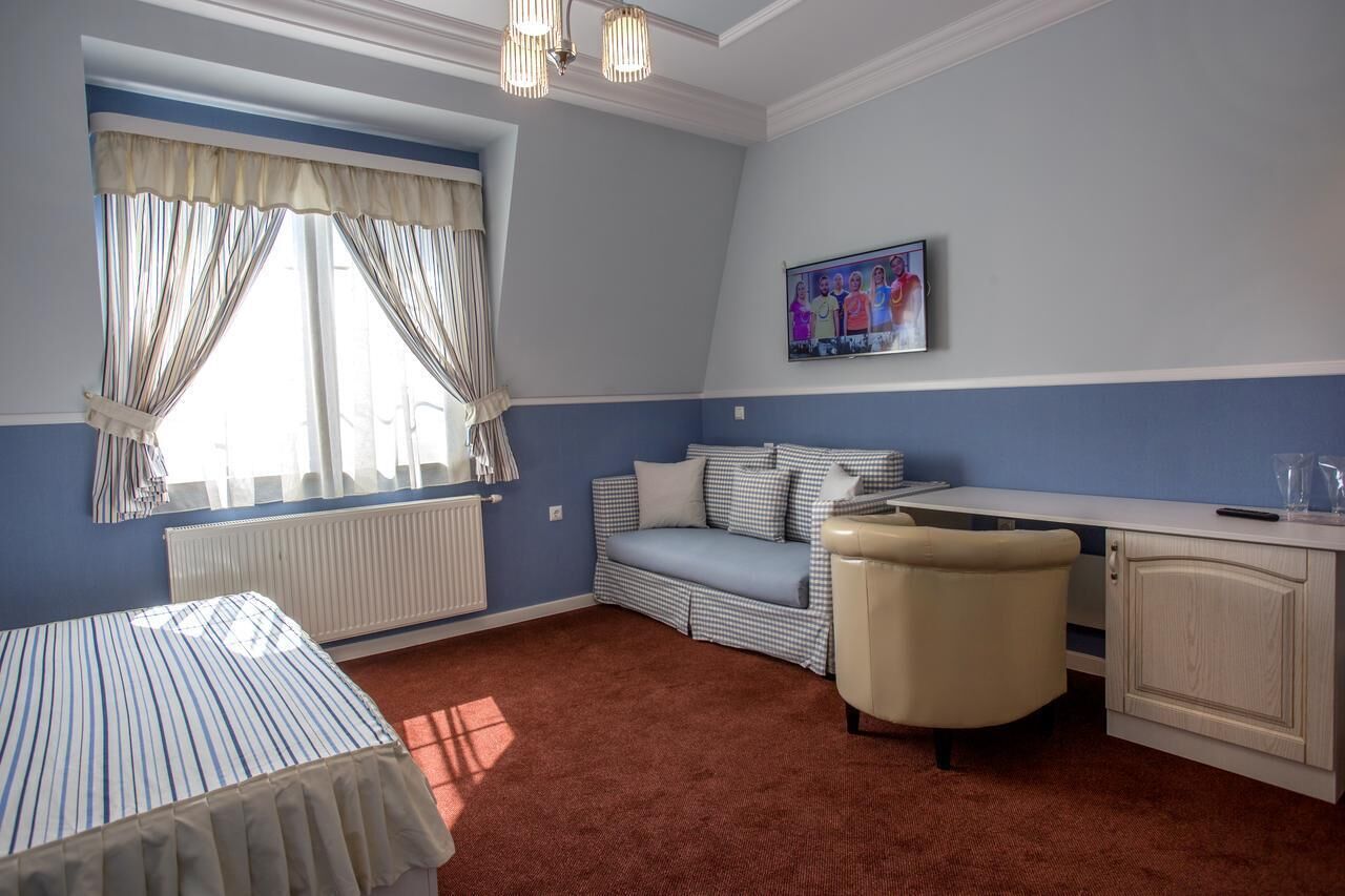 Una habitación con una cama a rayas, un sofá azul y blanco a cuadros, un sillón color beige, un escritorio blanco y un radiador.