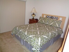 Een slaapkamer met een bed, nachtkastje, lamp en een raam met jaloezieën.