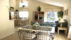 Zona pranzo con tavolo e sedie, un divano, televisione e lampada.