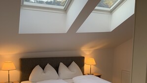 Een slaapkamer met een bed, twee bedlampen, een nachtkastje, een houten vloer en dakramen met uitzicht op bergen.