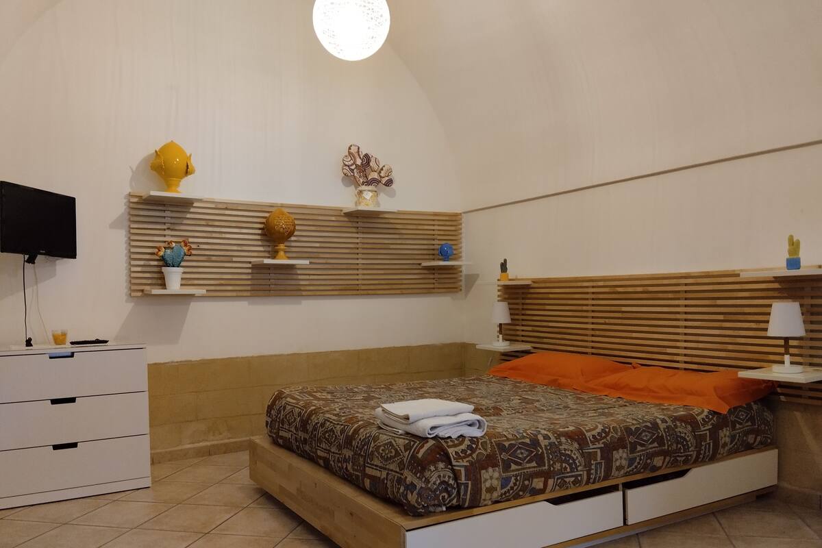 A bedroom with a bed, a TV, a wooden headboard, a bedside table, and a shelf with decorative items.