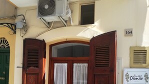 Una entrada de edificio con puerta de madera, dos ventanas con cortinas y una unidad de aire acondicionado encima, además de un letrero en la pared.