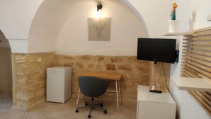 Una habitación con pared de piedra, un escritorio blanco, una silla, un pequeño armario blanco, un televisor montado en la pared y una estantería con una planta pequeña.