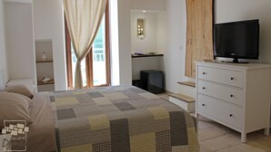 Un dormitorio con cama, televisor sobre un aparador, ventana con cortinas y un baño pequeño.