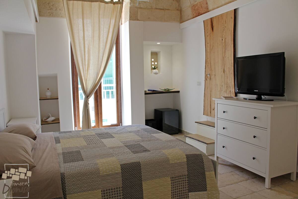 A bedroom with a bed, a TV on a dresser, a window with curtains, and a small bathroom.
