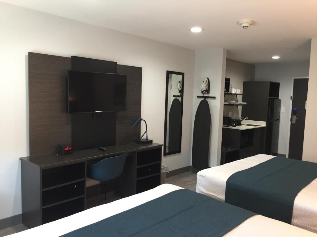 A hotel room with two beds, a desk, a chair, a TV, a mirror, and a kitchenette.