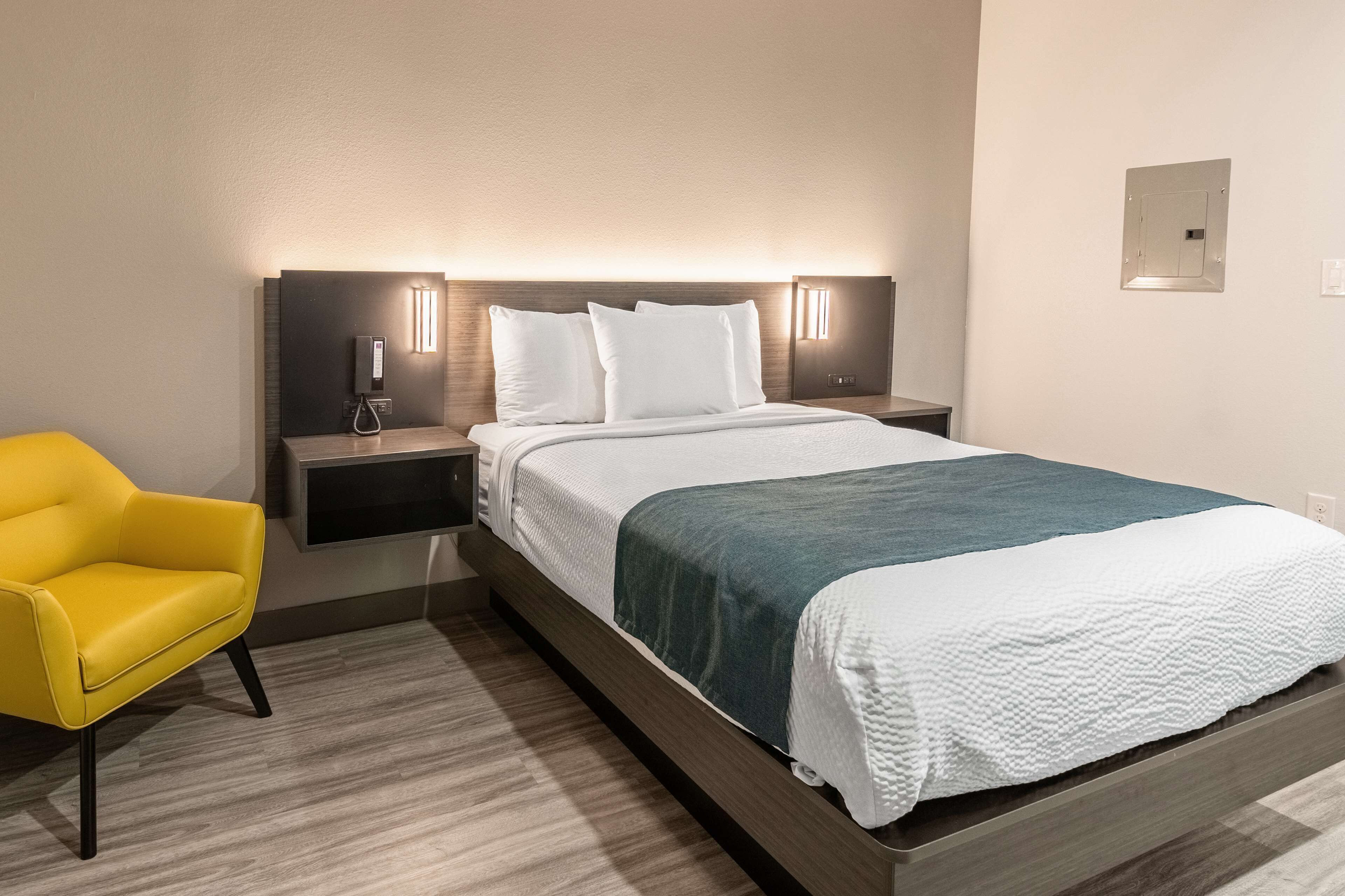 Une chambre d’hôtel avec un grand lit, une table de chevet avec un téléphone et une lampe, une chaise jaune et un luminaire fixé au mur.