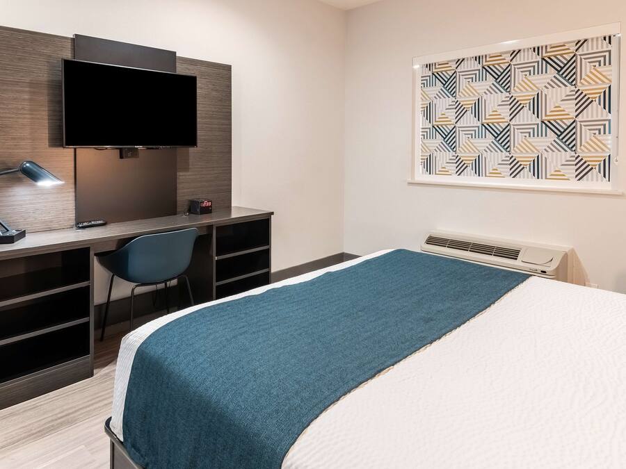 A hotel room with a bed, a desk with a TV, a chair, and a wall-mounted air conditioner.