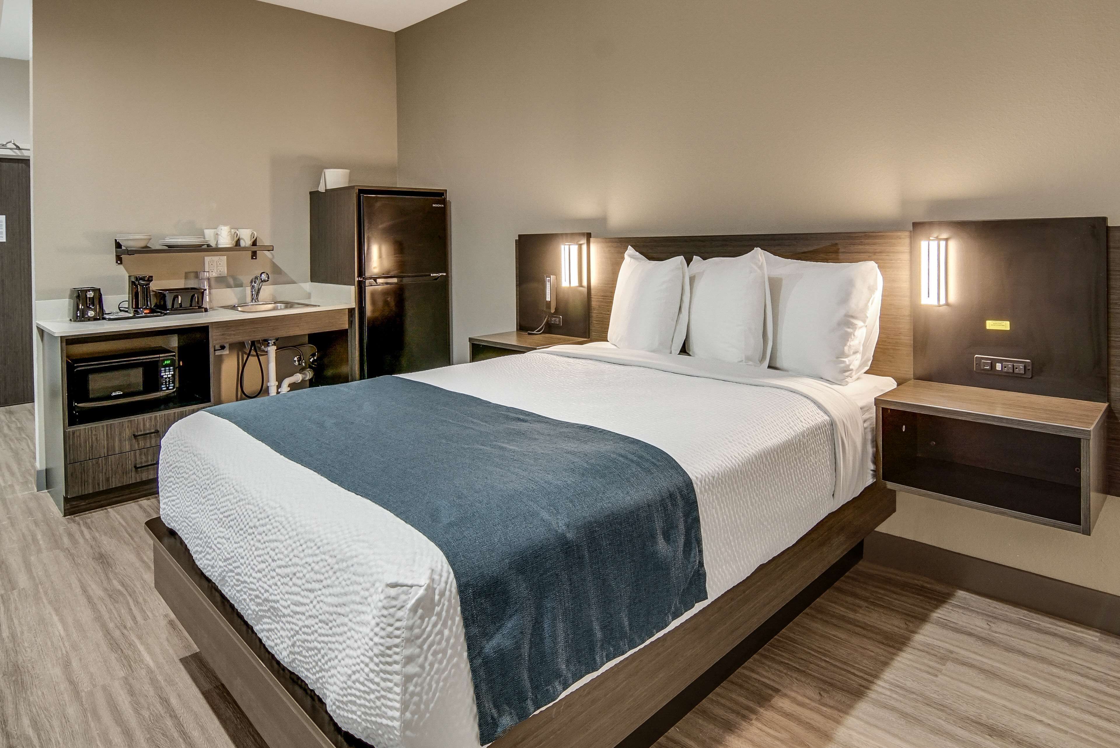 Habitación de hotel con cama, mesitas de noche, horno empotrado, refrigerador, pequeña cocina y teléfono fijo en la pared.