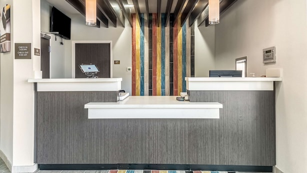 Un espace d’accueil d’hôtel au design moderne, comprenant un comptoir d’accueil en bois, un kiosque numérique pour l’enregistrement et un mur vertical aux couleurs vives.