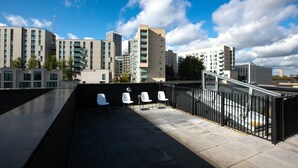 A rooftop terrace with a black railing, white chairs, and a view of surrounding buildings.