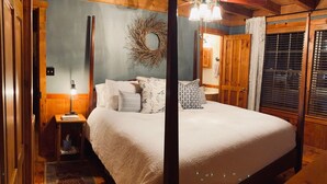 Une chambre confortable avec un plafond en bois, un lit recouvert d’une literie blanche, une table de chevet avec une lampe et une couronne décorative accrochée au mur.