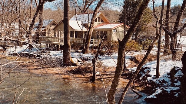 Une cabane rustique dotée d’une terrasse en bois et d’une rambarde en métal, entourée d’un paysage enneigé et d’arbres.