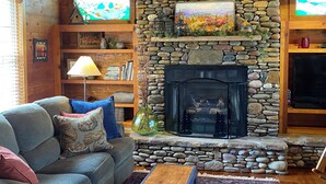 Un intérieur de chalet chaleureux avec une cheminée en pierre, des poutres en bois, un canapé avec des coussins, une bibliothèque et un téléviseur.