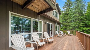 A wooden deck with Adirondack chairs, large windows, and a view of a forest. - Riesses Peace - Beautiful 4 bedroom, 3 bath home on Cupsuptic Lake (Oquossoc)