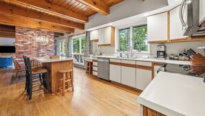 A modern kitchen with wooden beams, a brick accent wall, and a large island with bar stools. - Riesses Peace - Beautiful 4 bedroom, 3 bath home on Cupsuptic Lake (Oquossoc)