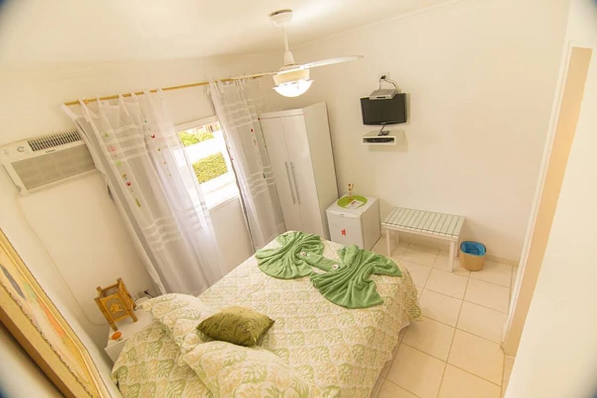 A bedroom with a bed, a bedside table, a small desk, a chair, a window with curtains, a ceiling fan, and a wall-mounted TV.