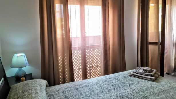 A hotel room with a bed, a nightstand with a lamp, and a window with curtains. - B&B Libellula (Iglesias)