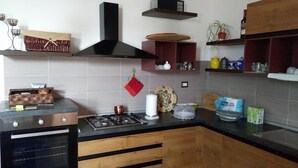 A kitchen with a gas stove, oven, and wooden cabinets. There are shelves with various kitchen items and a potted plant on the wall. - B&B Libellula (Iglesias)