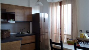 A compact kitchen with wooden cabinets, a stainless steel refrigerator, a sink, and a dining area with chairs and a table. - B&B Libellula (Iglesias)