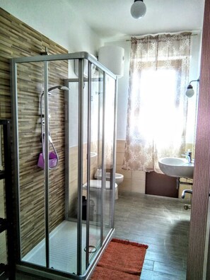 A bathroom with a glass shower enclosure, a toilet, a sink, and a window with curtains. - B&B Libellula (Iglesias)