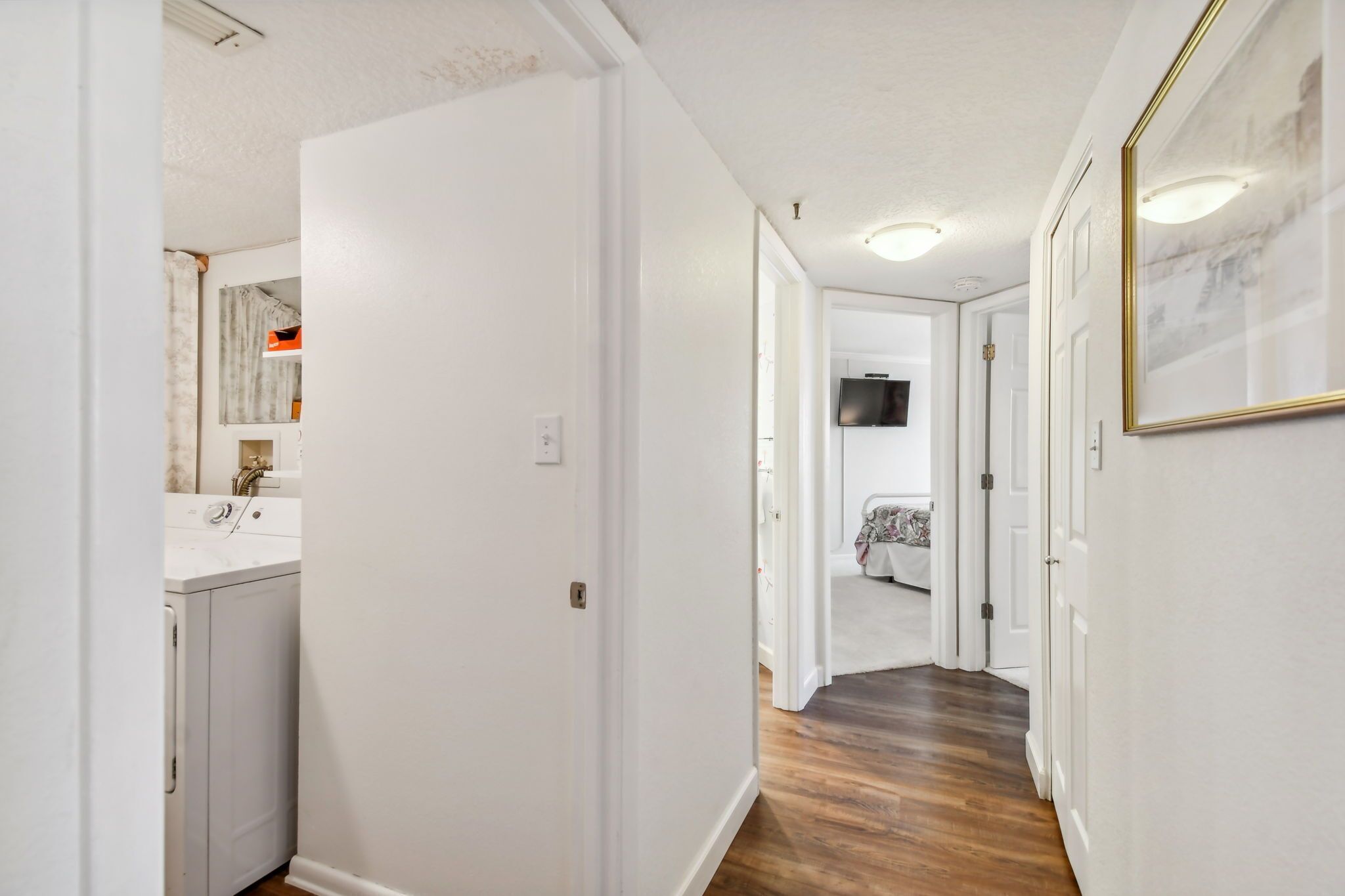 A hallway with a washing machine, a bed, and a television in a bedroom.