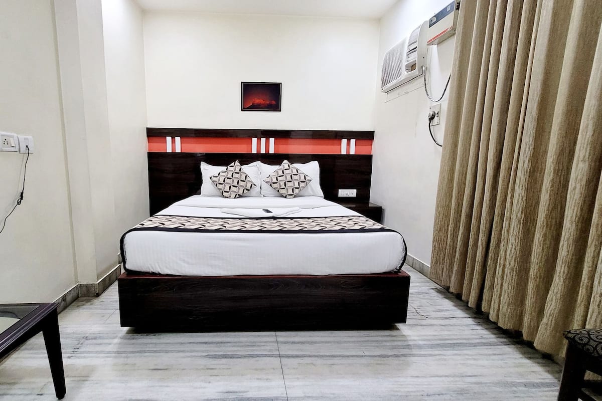 A neatly arranged hotel room with a bed, a bedside table, a power outlet, and a wall-mounted air conditioner.
